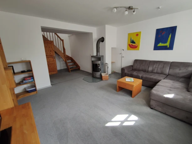 Heller Wohnbereich mit grauem Sofa, Holzofen, Treppe und modernen Kunstbildern an weißen Wänden.