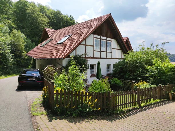 Zweistöckiges Fachwerkhaus mit roten Ziegeldach, umgeben von Wald, grüner Garten und parkendem Auto.