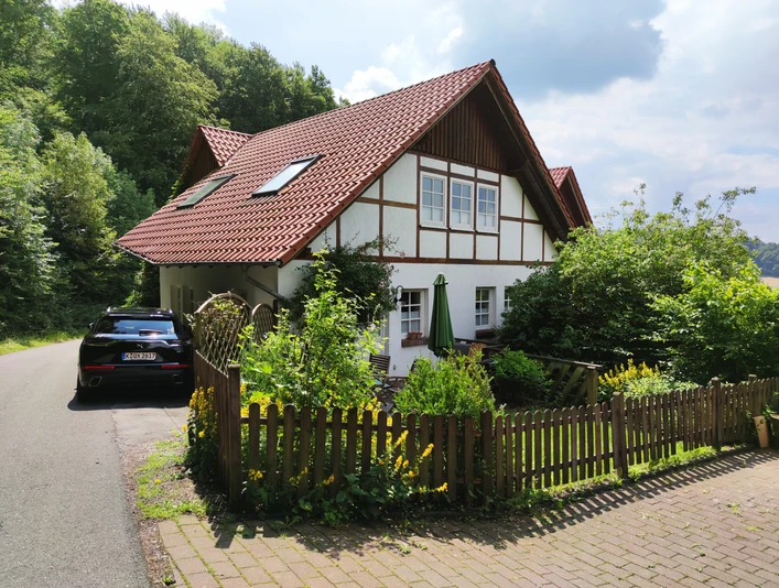 Haus am Wald.jpg Zweistöckiges Fachwerkhaus mit roten Ziegeldach, umgeben von Wald, grüner Garten und parkendem Auto.