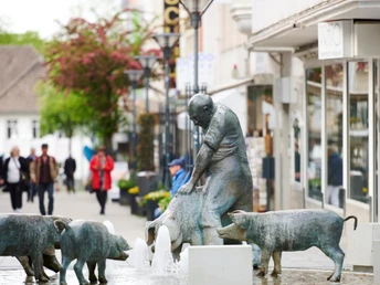 Bronzene Schweineskulpturen umrahmen einen Brunnen in einer belebten Fußgängerzone mit Geschäften.