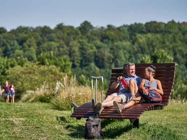 Rast am Knippeblick Zwei Personen sitzen entspannt auf einer Holzbank in grüner Landschaft, im Hintergrund wandern Leute.