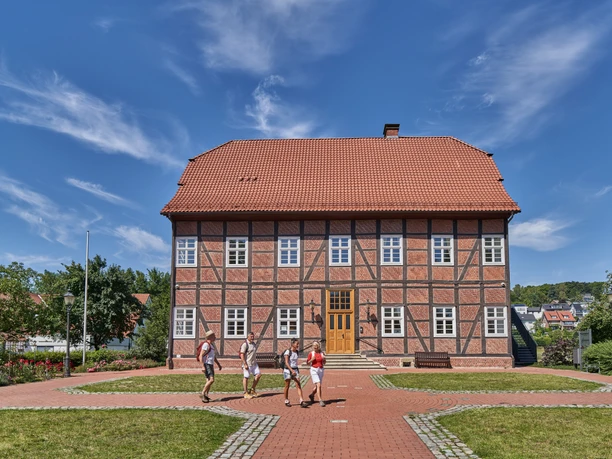Wandern am Spanckenhof Vier Personen spazieren vor einem beeindruckenden Fachwerkhaus mit rotem Dach und blauen Himmel.