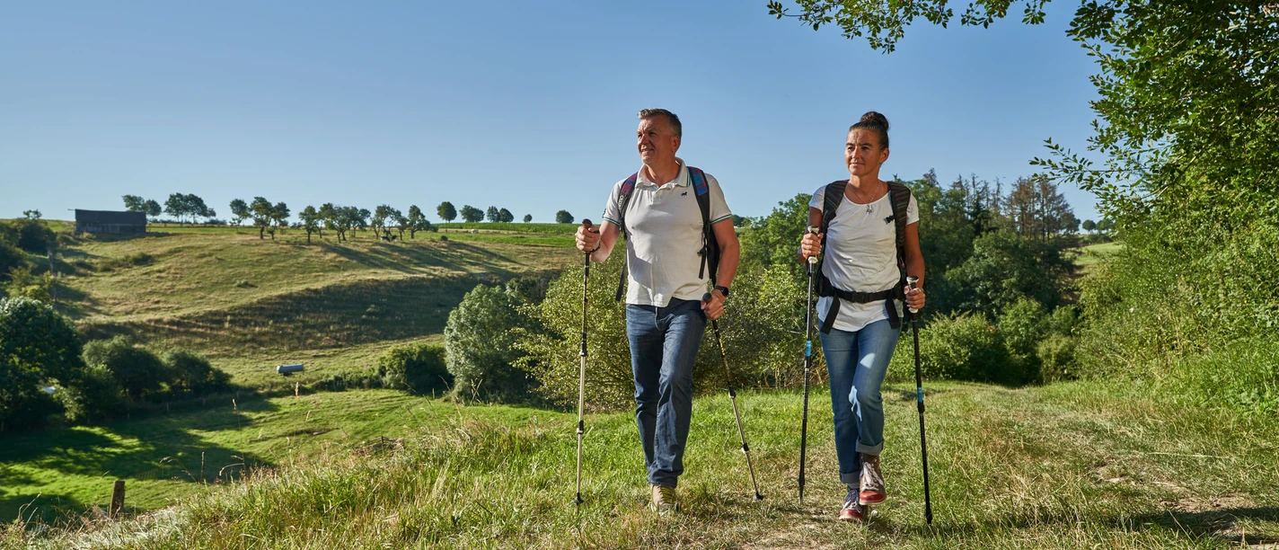 Wandern im Golmeketal Zwei Wanderer mit Stöcken gehen über eine grüne Wiese, Bäume und Hügel im Hintergrund.