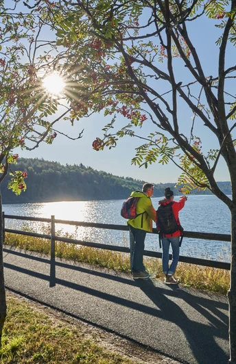 Zwei Wanderer genießen den Sonnenaufgang am Aabach, umgeben von Bäumen und ruhigem Wasser.