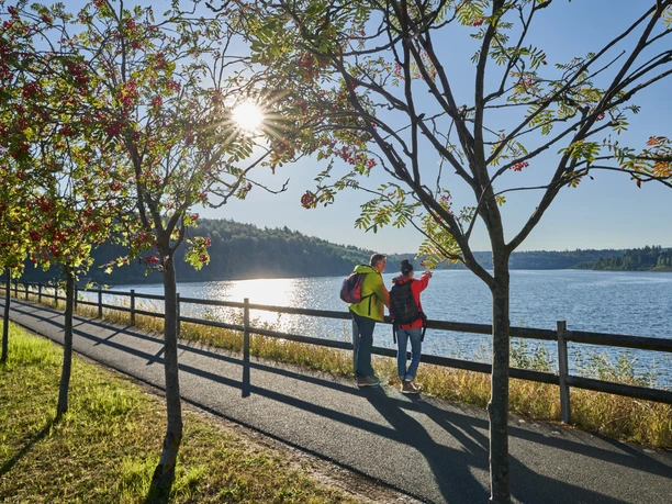 Sonnenaufgang an der Aabach-Talsperre Zwei Wanderer genießen den Sonnenaufgang am Aabach, umgeben von Bäumen und ruhigem Wasser.
