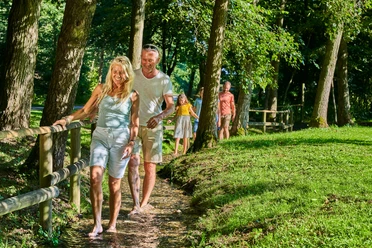 Personen genießen einen naturbelassenen Barfußpfad, umgeben von Bäumen und sommerlichem Grün.