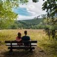 Ausblick auf die Schützenwiese Ein Paar sitzt auf einer Bank vor einer weiten Wiese mit Wäldern im Hintergrund, unter wolkigem Himmel.