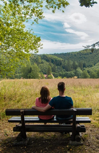 Ausblick auf die Schützenwiese Ein Paar sitzt auf einer Bank vor einer weiten Wiese mit Wäldern im Hintergrund, unter wolkigem Himmel.