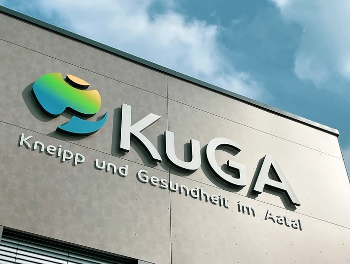 KuGA Fassade des Gesundheitszentrums KuGA mit modernem Logo und Schriftzug unter blauem Himmel.