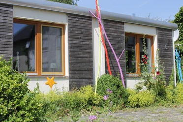 Außenansicht Flaches Gebäude mit Holzfassade, bunten Stangen und Pflanzen im Vordergrund, sonniges Wetter.