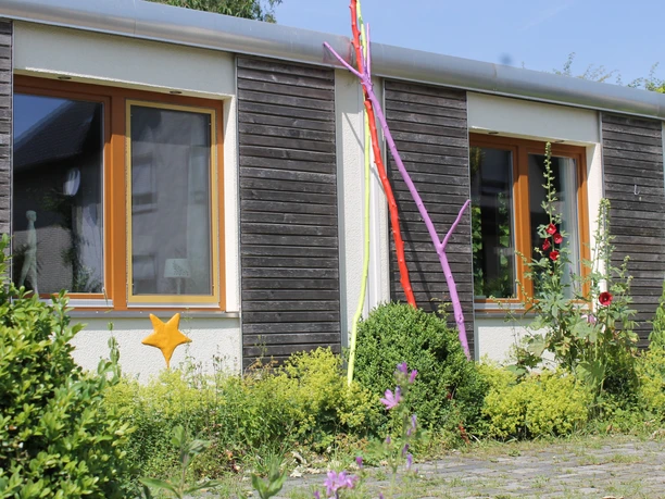 Außenansicht Flaches Gebäude mit Holzfassade, bunten Stangen und Pflanzen im Vordergrund, sonniges Wetter.