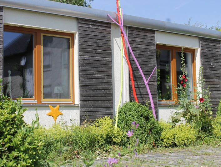 Außenansicht Flaches Gebäude mit Holzfassade, bunten Stangen und Pflanzen im Vordergrund, sonniges Wetter.