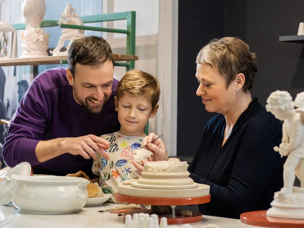 Erlebnis Schauwerkstatt Ein Kind lernt Töpfern, assistiert von einem Erwachsenen in einer Töpferwerkstatt.