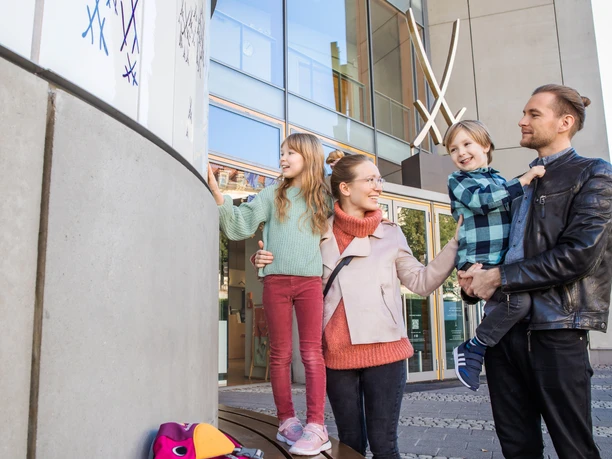 Erlebniswelt MEISSEN Ein fröhliche Familie mit zwei Kindern vor einem modernen Gebäude mit großen Glasfenstern.