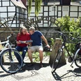 Zwei Radfahrer sitzen auf einer Bank vor historischem Fachwerkhaus und schauen gemeinsam in eine Karte.