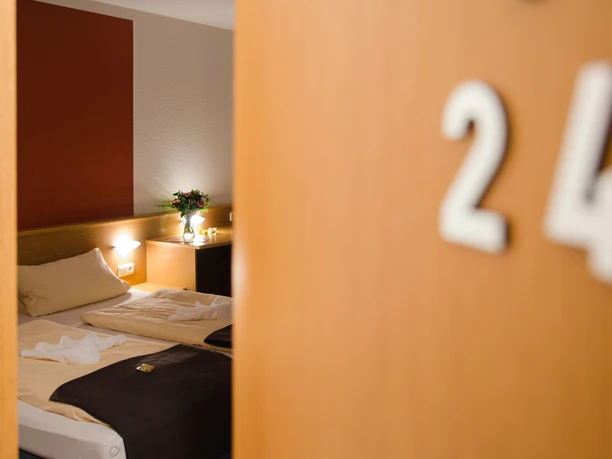 Landhaus Begatal Zimmertür CC BY-SA - Büker.jpg Geöffnete Zimmertür 24 mit Blick auf Hotelbett und Nachttisch im Landhaus Begatal