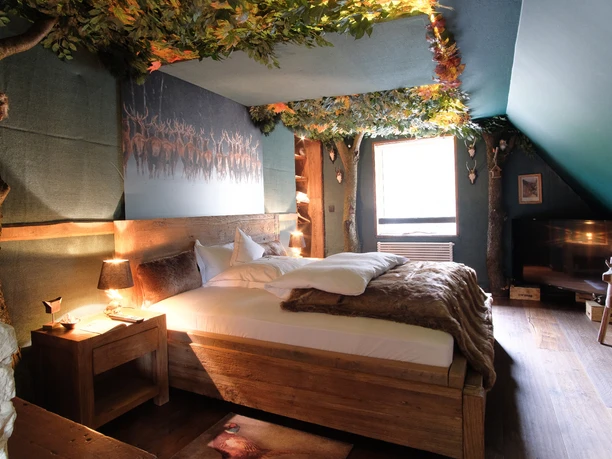 Suite Wald-Suite mit rustikalem Holzbett, Felldecke, Baumstämmen, Blätterdach und Hirschbild.