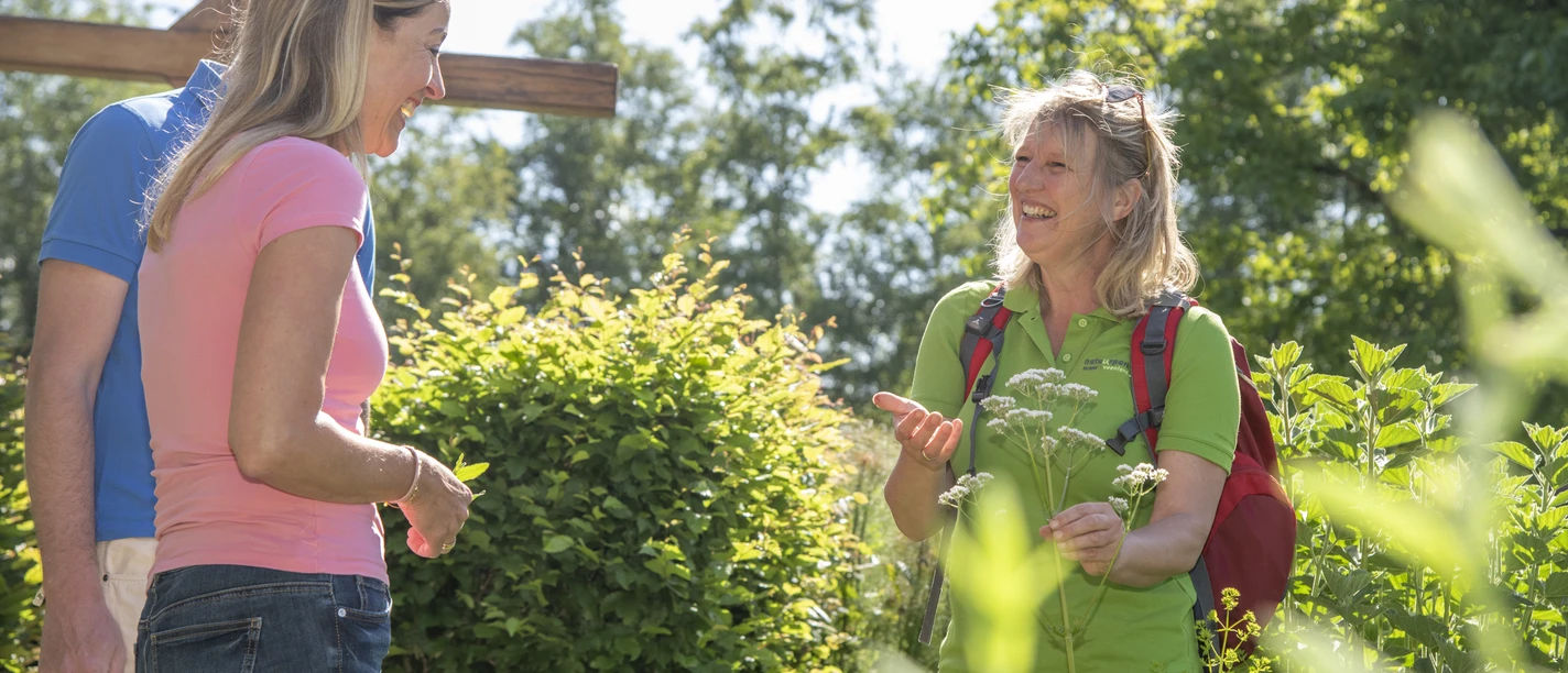 Bibelgarten Twist, Garten des Nazareners ©TMN GmbH, Sabine Braun Fotografie-31.jpg Zwei Frauen sprechen im sonnigen Garten zwischen grünen Pflanzen und einem hölzernen Kreuz.