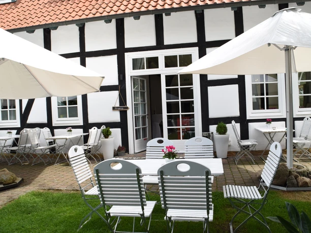 Gepflegte Terrasse mit weiß-schwarzem Fachwerkhaus, Tischen, Stühlen und Sonnenschirmen im Garten.