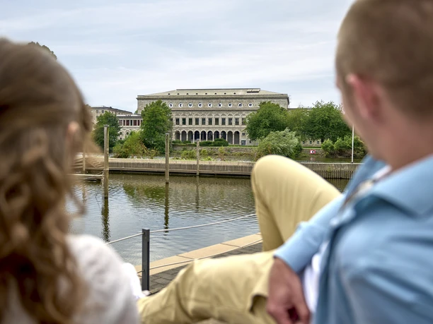 Stadthalle, Mülheim an der Ruhr Blick auf die Stadthalle Mülheim, ein imposantes Gebäude am Fluss mit Bäumen und Menschen im Vordergrund.