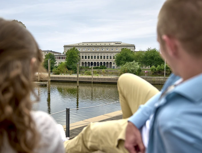 Stadthalle, Mülheim an der Ruhr Blick auf die Stadthalle Mülheim, ein imposantes Gebäude am Fluss mit Bäumen und Menschen im Vordergrund.