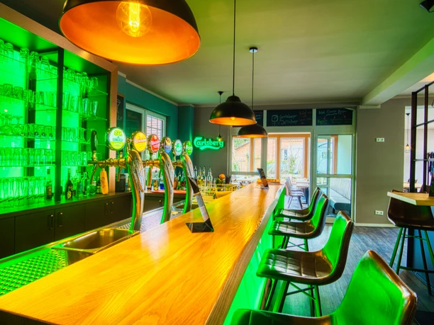 Innenansicht Moderne Bar mit langer Holzteke, grüner LED-Beleuchtung und schwarzen Barhockern.