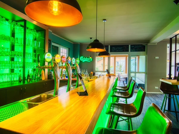 Innenansicht Moderne Bar mit langer Holzteke, grüner LED-Beleuchtung und schwarzen Barhockern.