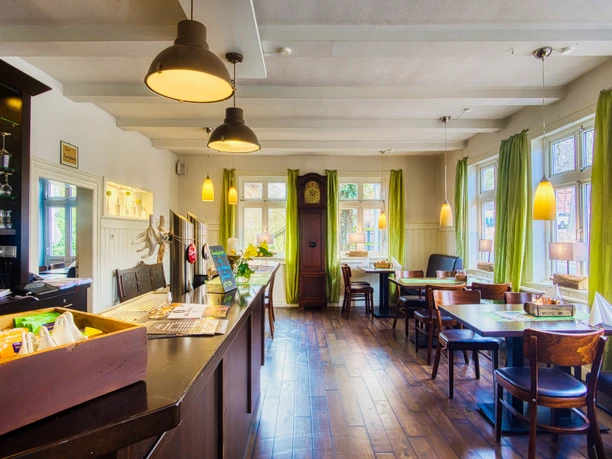 Gastraum Heller Gastraum mit dunklem Holzboden, Theke, Tischen und grünen Vorhängen an den Sprossenfenstern.