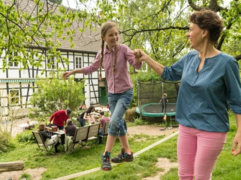 Freizeit auf Hof zur Hellen bei Velbert-Neviges Mutter und Tochter balancieren fröhlich auf Seil, während im Hintergrund Menschen im Garten entspannen.