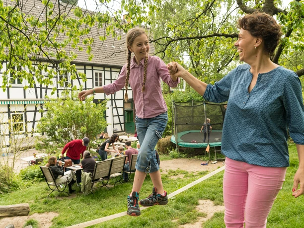 Freizeit auf Hof zur Hellen bei Velbert-Neviges Mutter und Tochter balancieren fröhlich auf Seil, während im Hintergrund Menschen im Garten entspannen.