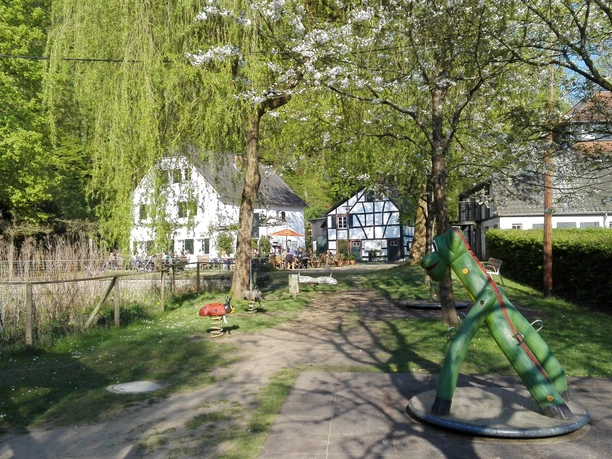 Alte Dombach Ein idyllischer Spielplatz mit Wippe und Fachwerkhäusern im Hintergrund unter blühenden Bäumen.