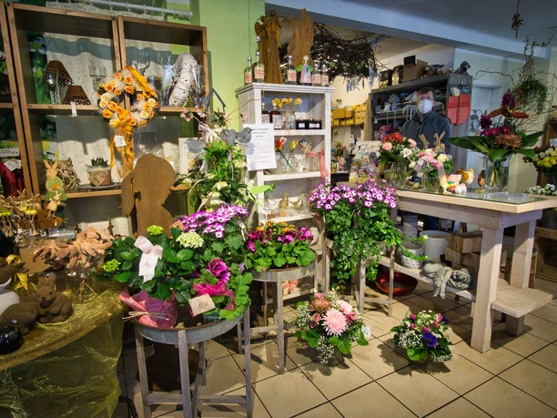 Blüte und Stil Innenaufnahme eines Blumenladens mit verschiedenen Pflanzenarrangements und handgefertigten Dekorationen. Mehrere Blumentöpfe stehen auf Holztischen, umgeben von floralen Kränzen und Pflanzenaccessoires, die eine warme Atmosphäre schaffen.