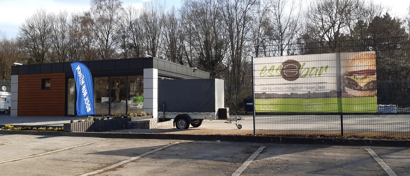Ein moderner, flacher Imbissstand mit Holzverkleidung und großer Werbetafel, umgeben von Bäumen.