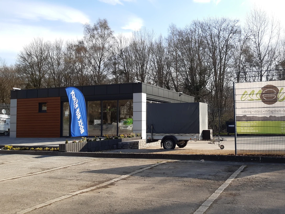 Essbar | Büren Ein moderner, flacher Imbissstand mit Holzverkleidung und großer Werbetafel, umgeben von Bäumen.