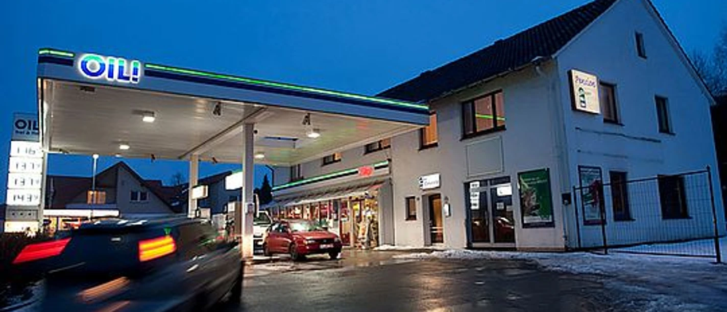 OIL-INN.jpg Eine Tankstelle im Abendlicht mit mehreren Zapfsäulen, daneben ein Gebäude aus weißem Putz.