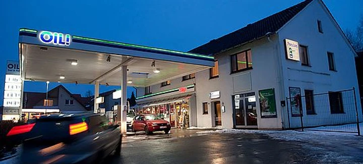 OIL-INN.jpg Eine Tankstelle im Abendlicht mit mehreren Zapfsäulen, daneben ein Gebäude aus weißem Putz.