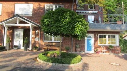 Apartment Bärenhöhle.JPG Rotes Backsteinhaus mit blauen und weißen Türen, gepflegtem Vorgarten und Baum in der Mitte.