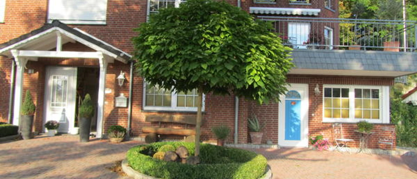 Apartment Bärenhöhle.JPG Rotes Backsteinhaus mit blauen und weißen Türen, gepflegtem Vorgarten und Baum in der Mitte.