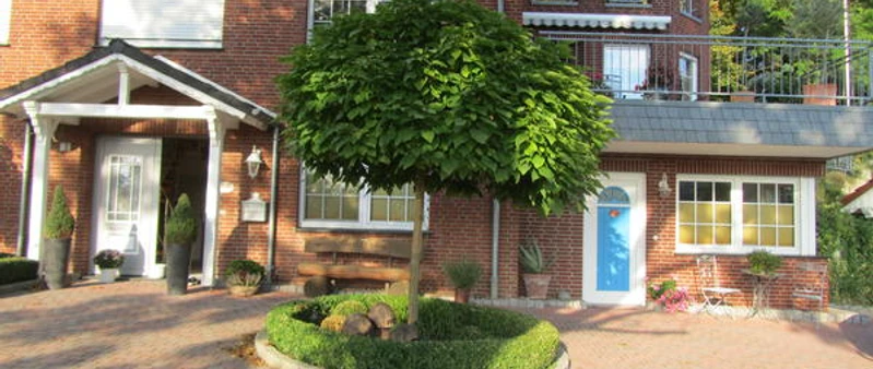 Apartment Bärenhöhle.JPG Rotes Backsteinhaus mit blauen und weißen Türen, gepflegtem Vorgarten und Baum in der Mitte.