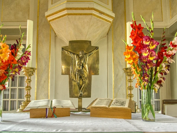Altar Ein goldener Altar mit einem bronzenen Christus im Zentrum, flankiert von bunten Gladiolen und zwei geöffneten Bibeln.