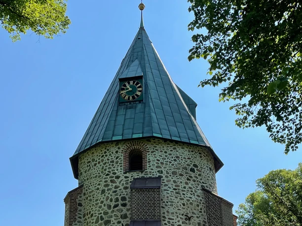 Kirche Betzendorf Runder Kirchturm aus Feldsteinen mit grünem Spitzdach, Uhr und Wetterfahne unter blauem Himmel.
