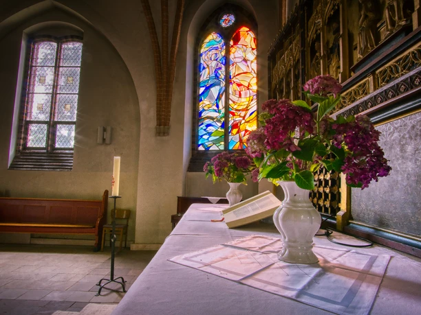 Innenansicht Altarraum der St. Peter und Paul Kirche mit buntem Glasfenster und lila Hortensien.