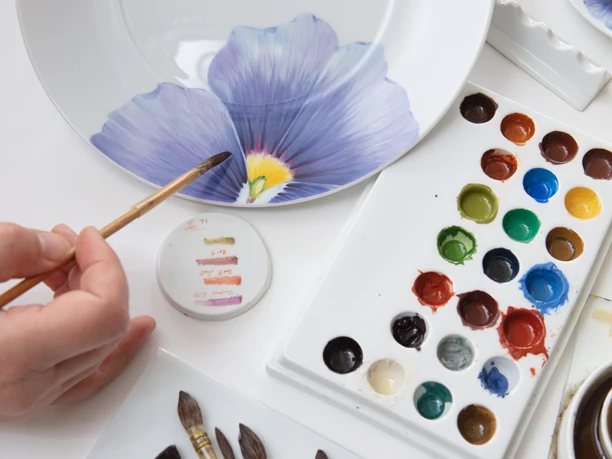 Aufglasur Giant Bloom Person bemalt Porzellanteller mit blauer Blume, umgeben von Pinseln und Farbpalette.