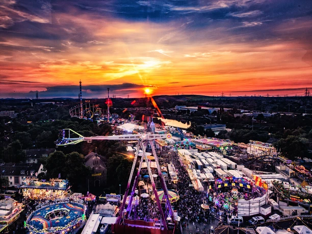 Luftaufnahme eines bunten Volksfests bei Sonnenuntergang mit Karussells und vielen Besucher*innen.