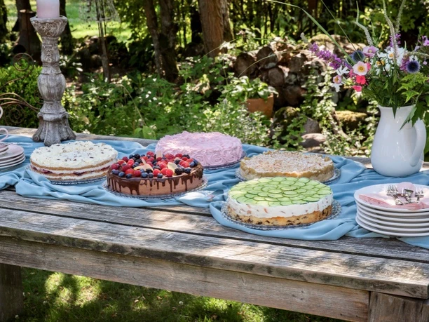 Buntes Tortenbuffet auf einem Holztisch im grünen Garten mit Blumen und Geschirr.
