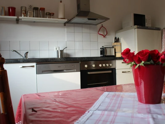 Ferienwohnung "Numero 7" Wohnküche mit rotem Tisch, Vasenrosen, moderner Küche und hellem Keramikfliesenrückwand.