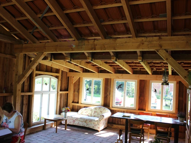 Melkhuske_Schatteburg_MKul_22.8 (72).JPG Heller Holzraum im Melkhuske mit Balken, geblümtem Sofa, Tischen und einer sitzenden Frau.