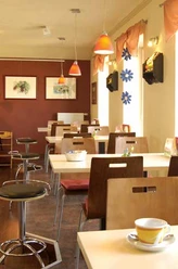 cafe vielfalt.jpg Ein helles Café mit modernen Holzmöbeln, orangefarbener Beleuchtung und Kunst an den Wänden.