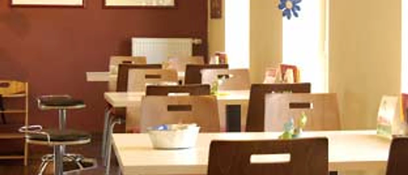 cafe vielfalt.jpg Ein helles Café mit modernen Holzmöbeln, orangefarbener Beleuchtung und Kunst an den Wänden.