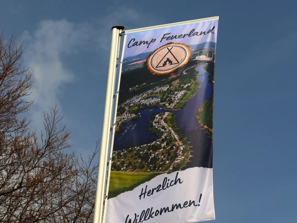 Fahne Feuerland Eine Fahne, auf der die Weser und der Campingplatz Feuerland abgebildet sind. Auf der Fahne steht "Camp Feuerland Herzlich Willkommen"
